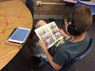 Boy sitting at a table. He has headphones on and is reading a graphic novel. 